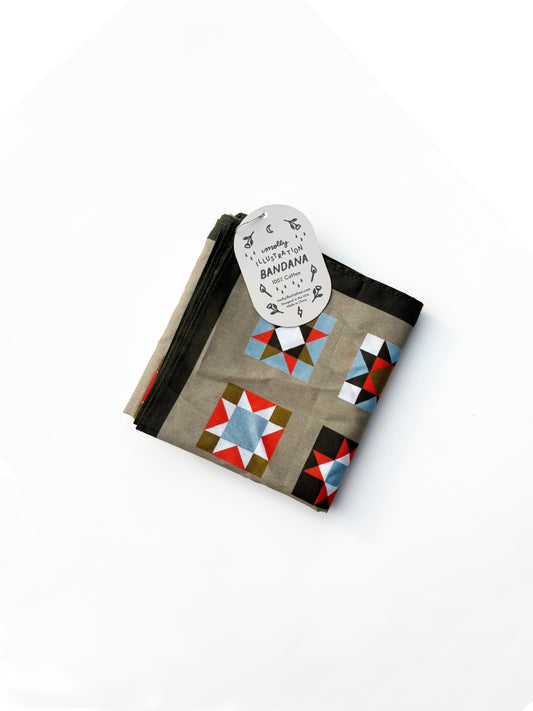 A folded cotton bandana featuring a geometric star quilt pattern in red, blue, white, and brown on a taupe background, with a black border and a white paper tag attached.