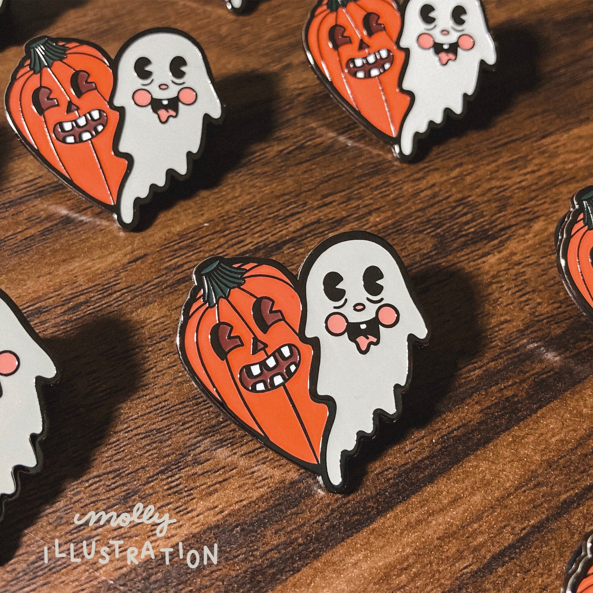 several illustrated hard enamel pins of a ghost and jack o lantern pumpkin over a wooden background. 