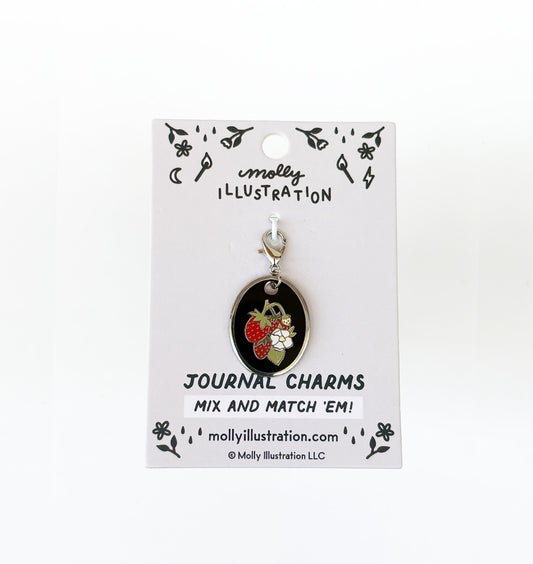 A silver, black, red and green  journal charm with strawberries on it. Behind it is a branded backer card for the product
