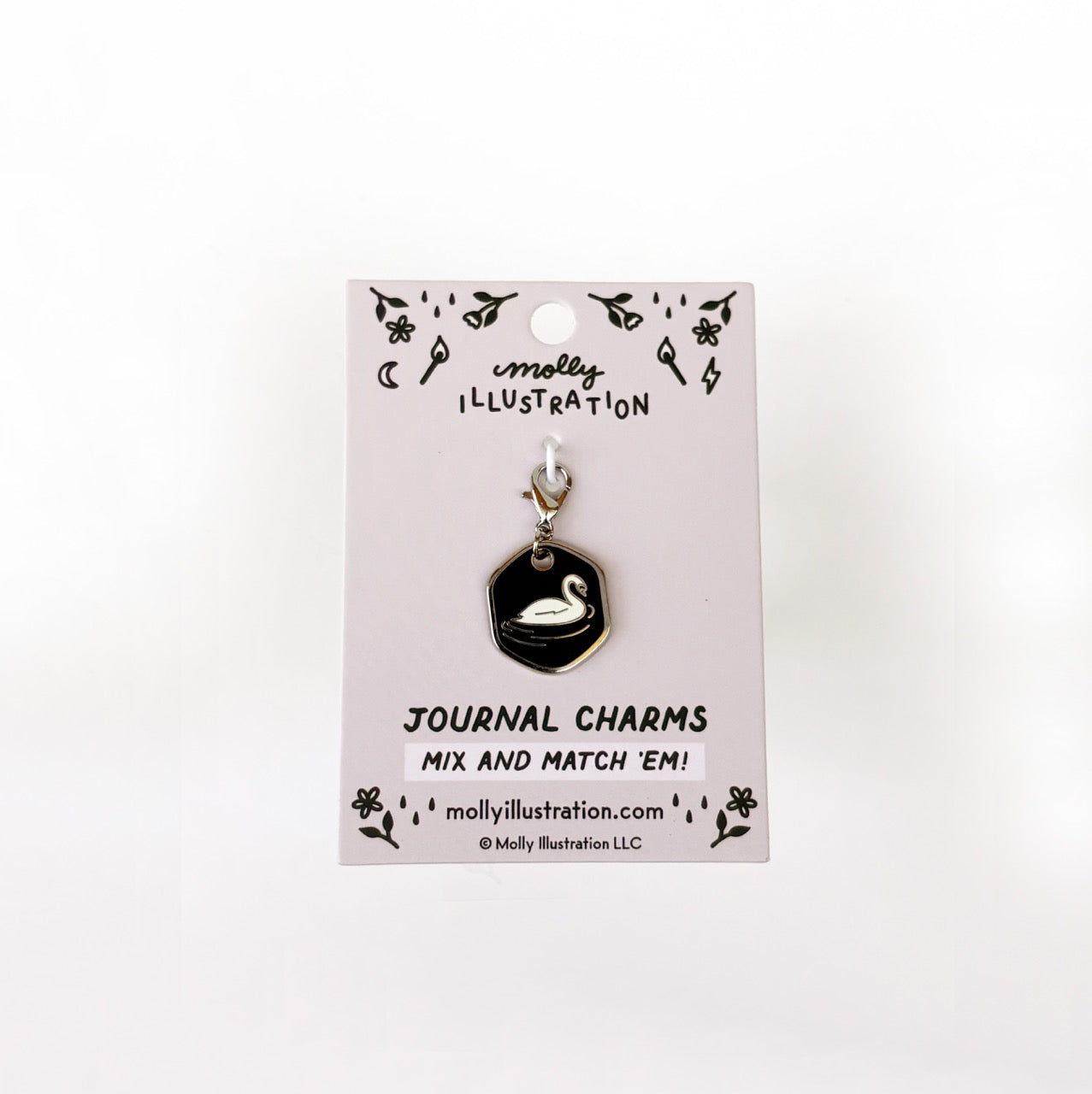 A silver and black journal charm with a swan on it. Behind it is a branded backer card for the product