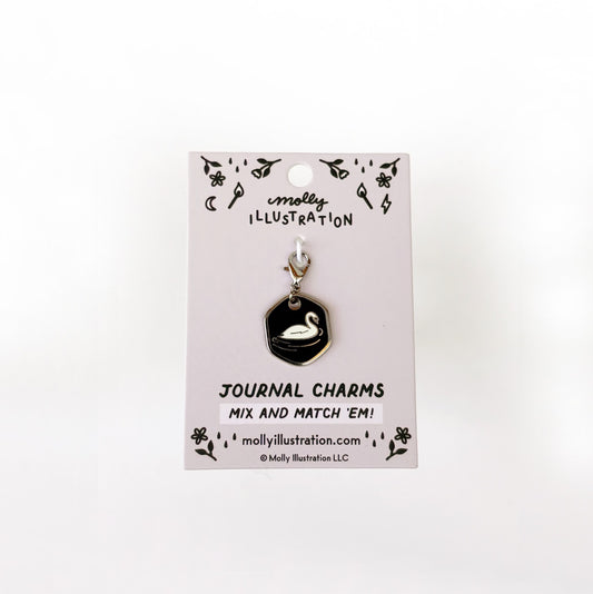 A silver and black journal charm with a swan on it. Behind it is a branded backer card for the product