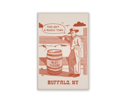 a vintage inspired illustrated magnet of a cartoon cowboy beside a barrel of bleu cheese. The text beside it reads "this ain't a ranch town." and Buffalo, NY below it.
Beside it is the back view of the magnet flipped over.