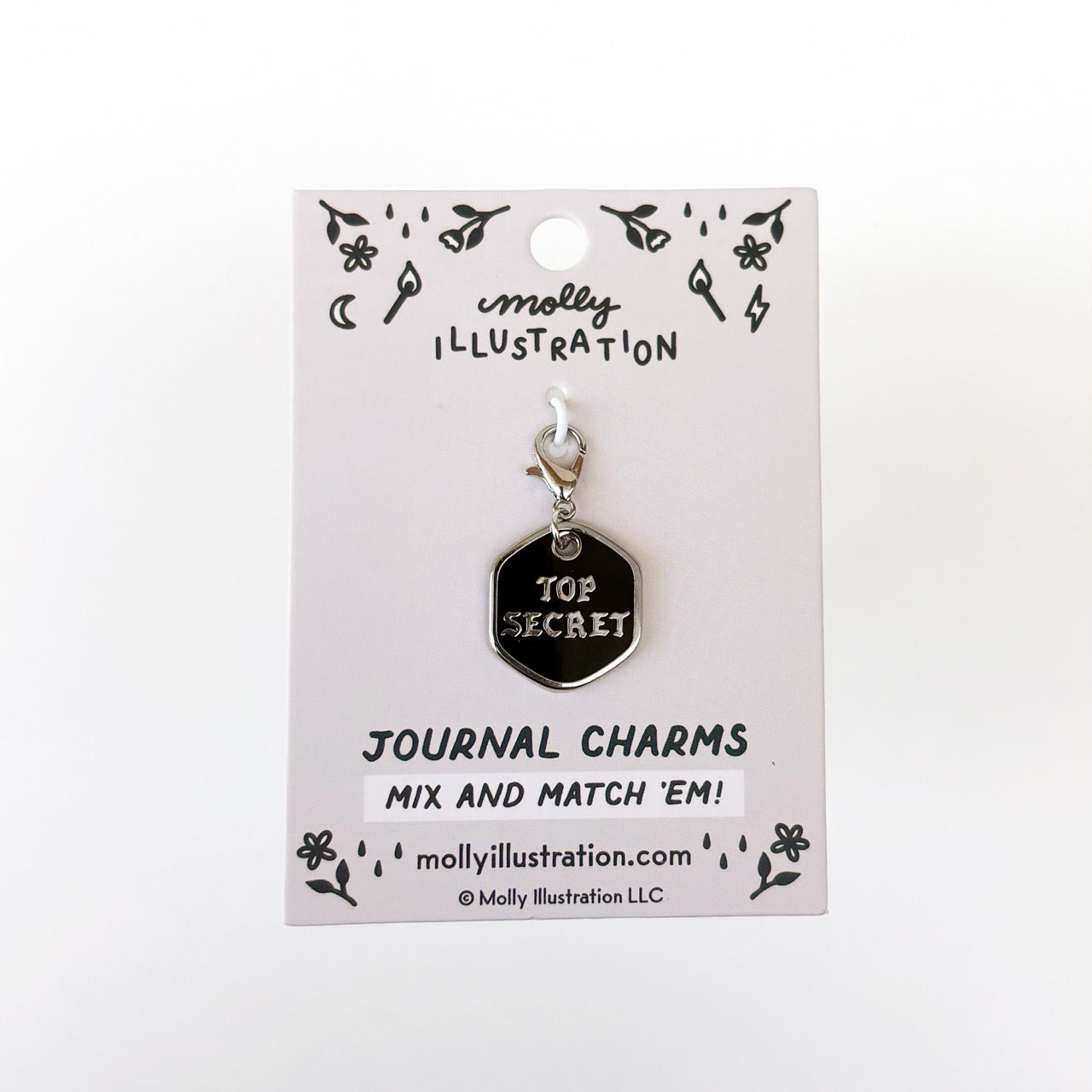 A silver and black journal charm with top secret on it. Behind it is a branded backer card for the product