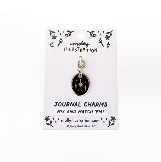 A silver and black journal charm with stars on it. Behind it is a branded backer card for the product