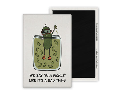 a white and green illustrated refrigerator magnet of a pickle swimming in a pickle jar that reads "We say in a pickle like it's a bad thing" Beside it is the back view of the magnet flipped over.