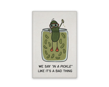 a white and green illustrated refrigerator magnet of a pickle swimming in a pickle jar that reads "We say in a pickle like it's a bad thing" Beside it is the back view of the magnet flipped over.