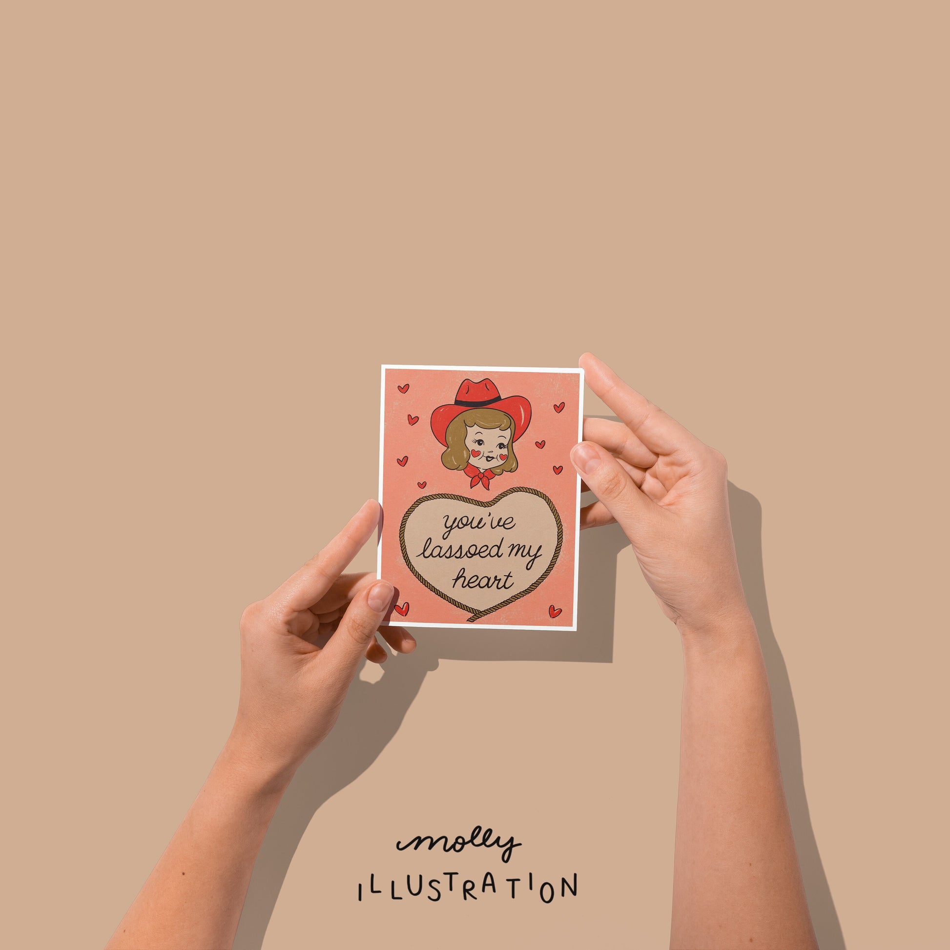 Hands holding an illustrated western greeting card that reads "you've lassoed my heart" surrounded by red hearts and a cute illustrated cowgirl above the text.
