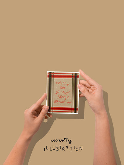 hands holding an illustrated greeting card that reads "wishing you a very merry christmas" with a plaid tan, red and green background.