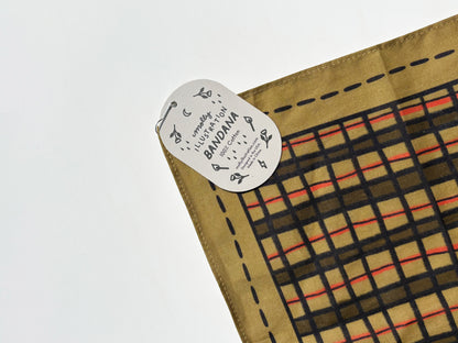 A close-up of a square woven textile corner with a white paper tag reading 'BANDANA' and 'handwoven cloth'. The fabric has a tan base with a black grid pattern and thin red lines, photographed on a plain white background.