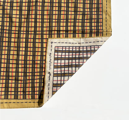 A folded patterned scarf  shown on a clean white background. The visible surface has a plaid-style grid in black, peach, orange, and beige, with a tan border and a folded corner revealing the reverse side and printed text along the edge.