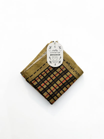A folded cotton bandana with a plaid pattern in black, beige, and orange-red, featuring a gold-brown border and a white paper hangtag.