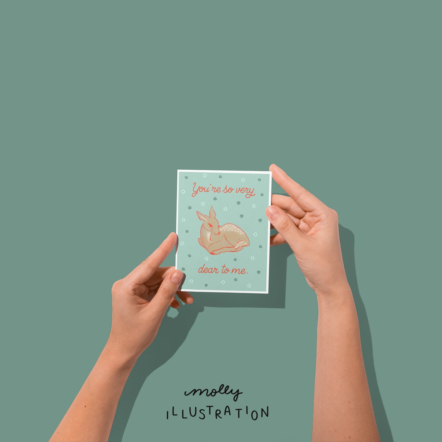 hands holding an illustrated greeting card that reads "you're so very dear to me" around a drawing of a brown deer with a blue background.