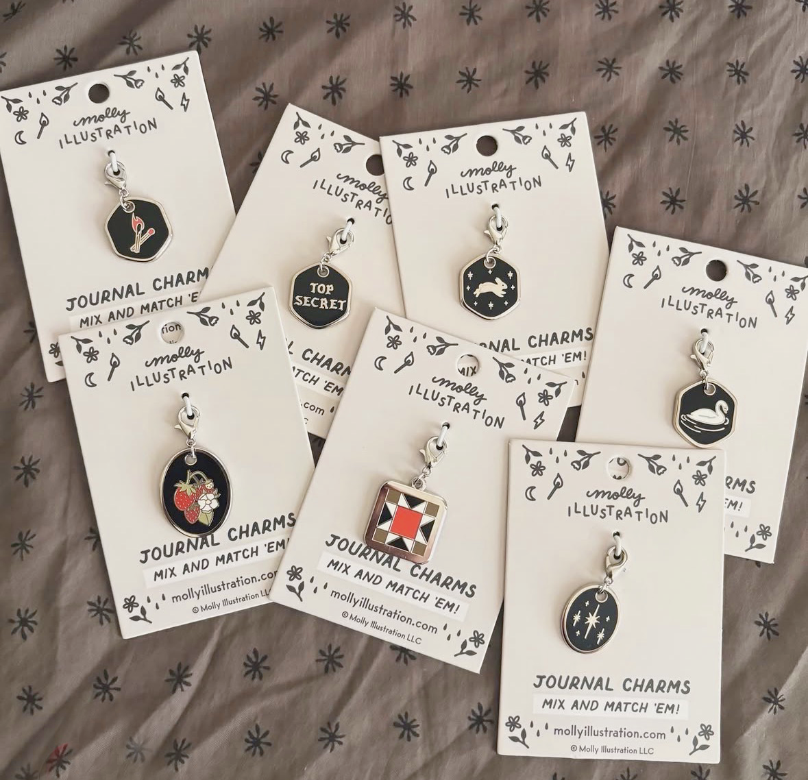 Seven journal charms displayed on white backing cards, arranged on a gray patterned fabric background. 